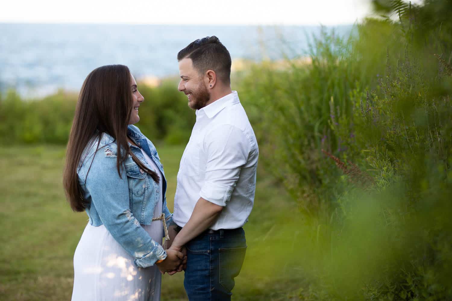 A man and woman stand outdoors facing each other, holding hands and smiling, with greenery and water in the background.