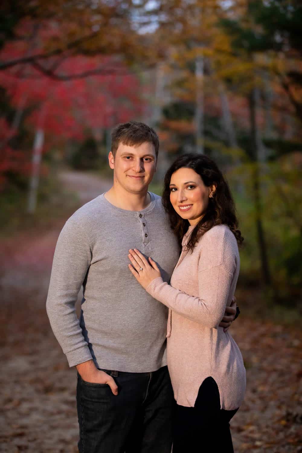 A couple stands closely together on a forest path with autumn foliage in the background, both smiling at the camera.