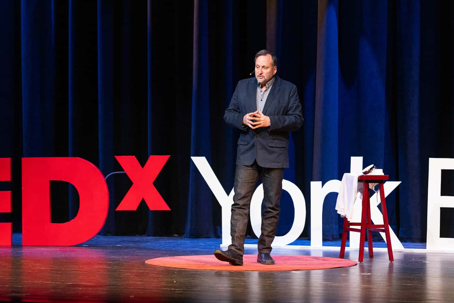 Man in a suit stands on a red circular carpet giving a talk on stage with large TEDx and other letters behind him and a small table with a cloth beside him.