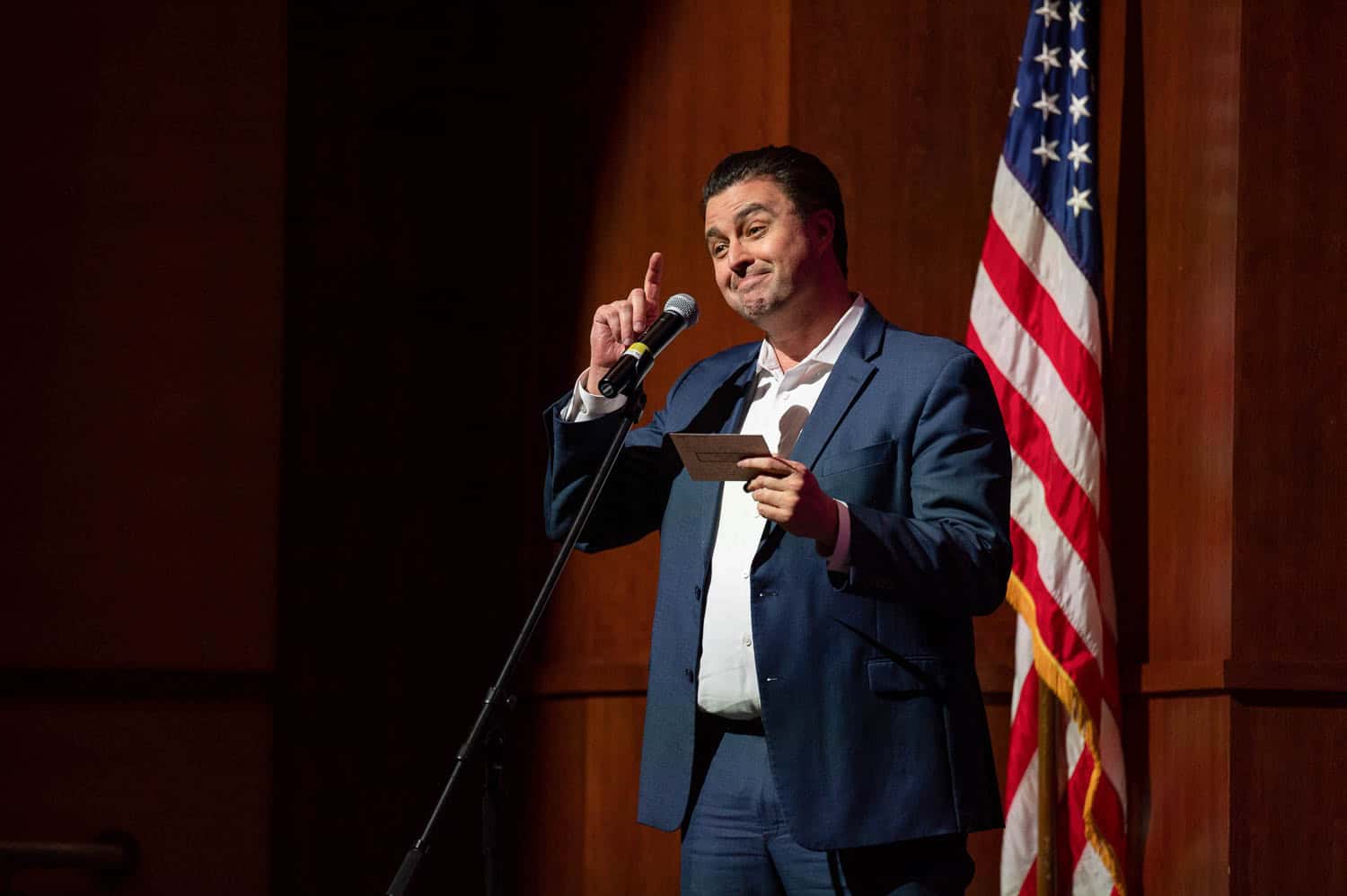 A man in a blue suit speaks into a microphone while holding a card, standing next to an American flag on a stage.