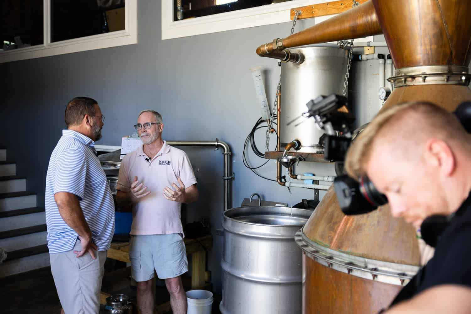 Two men converse in a distillery while a cameraman records nearby; metal tanks and copper equipment are visible in the background.