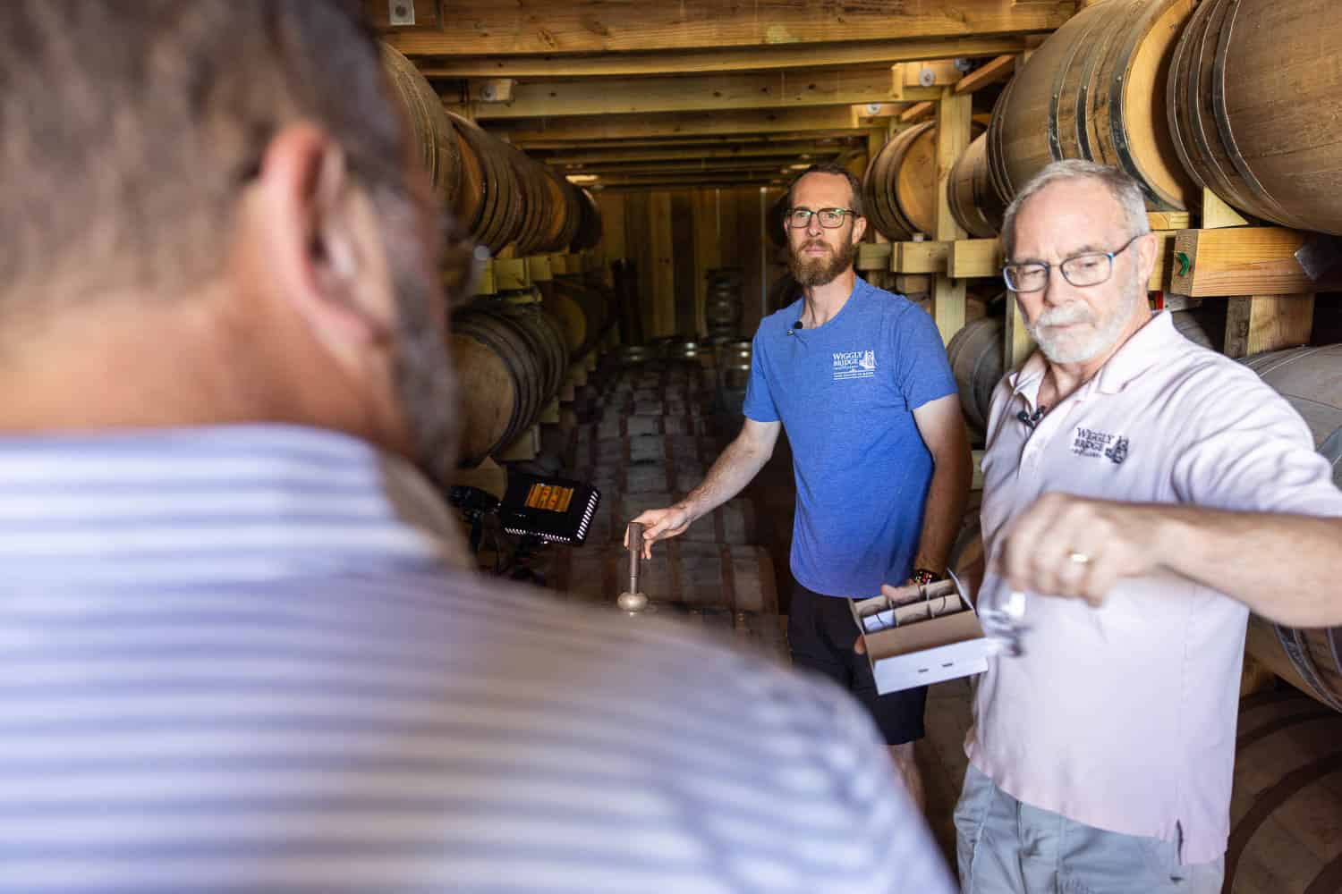 Two men stand in a barrel storage room, one holding a sample glass and the other holding a tray of glasses, while a third person is seen from behind.