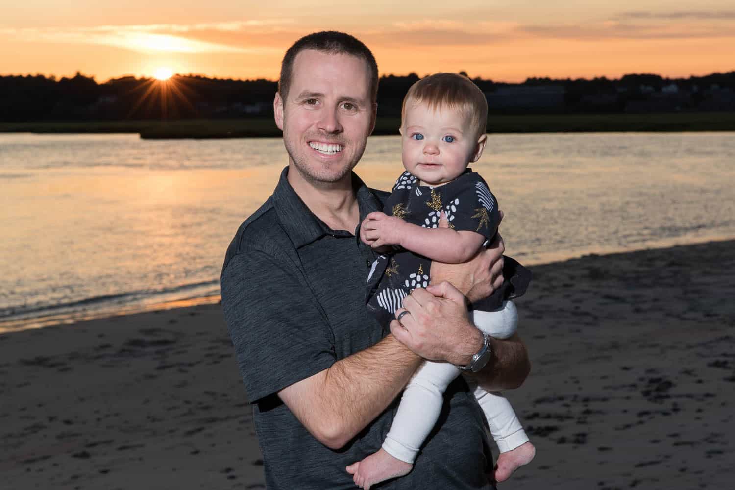 A man holds a baby while standing on a beach at sunset, with water and trees visible in the background.