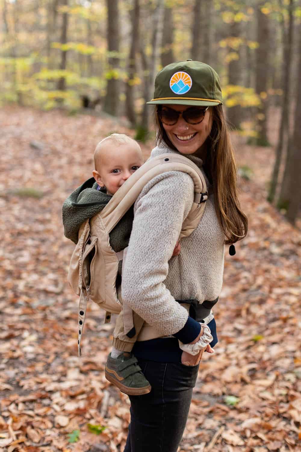 Woman wearing sunglasses and a cap carries a baby in a front-facing beige carrier while walking in a forest with fallen leaves on the ground.