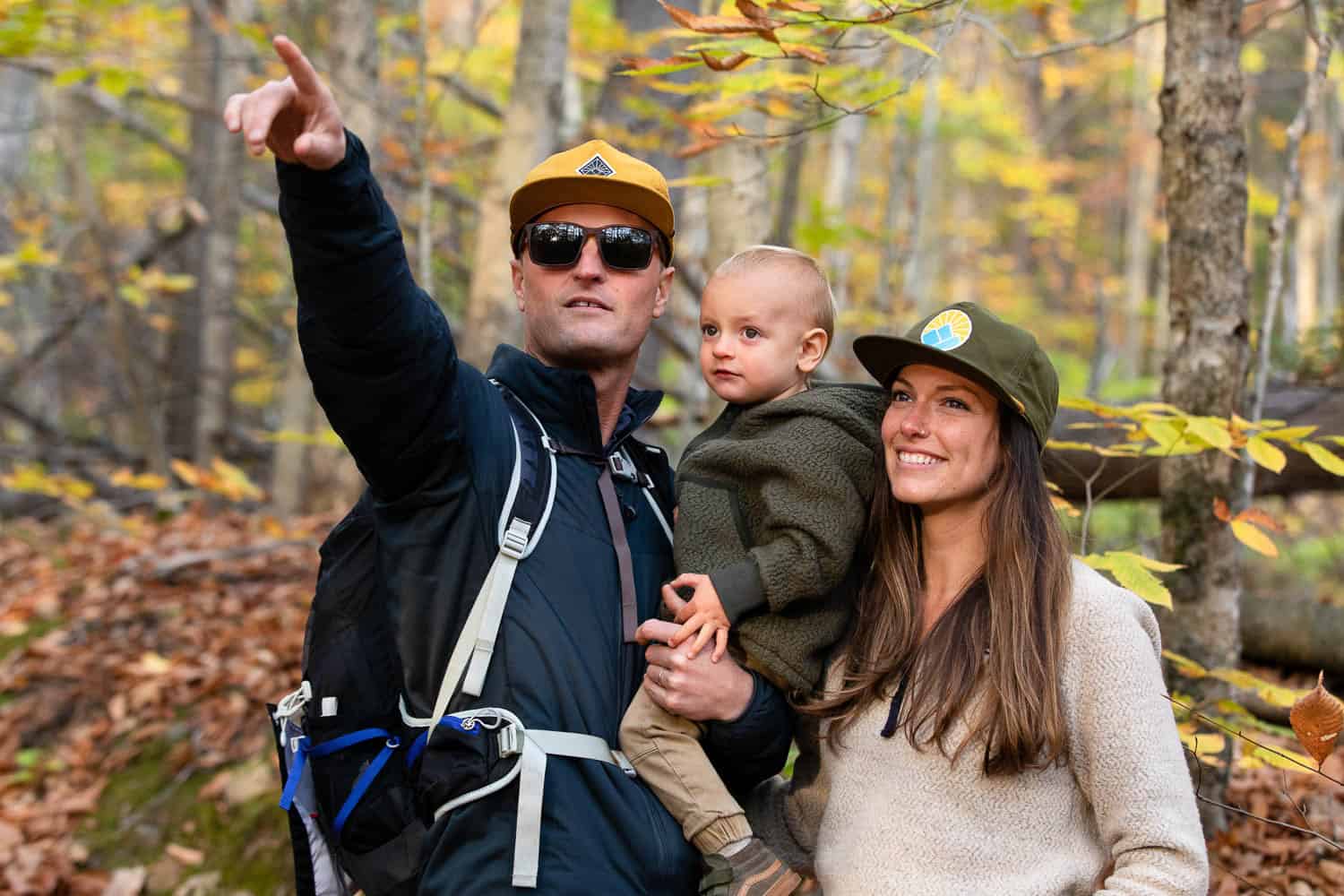 A man points while holding a young child next to a smiling woman in a forest with autumn leaves.