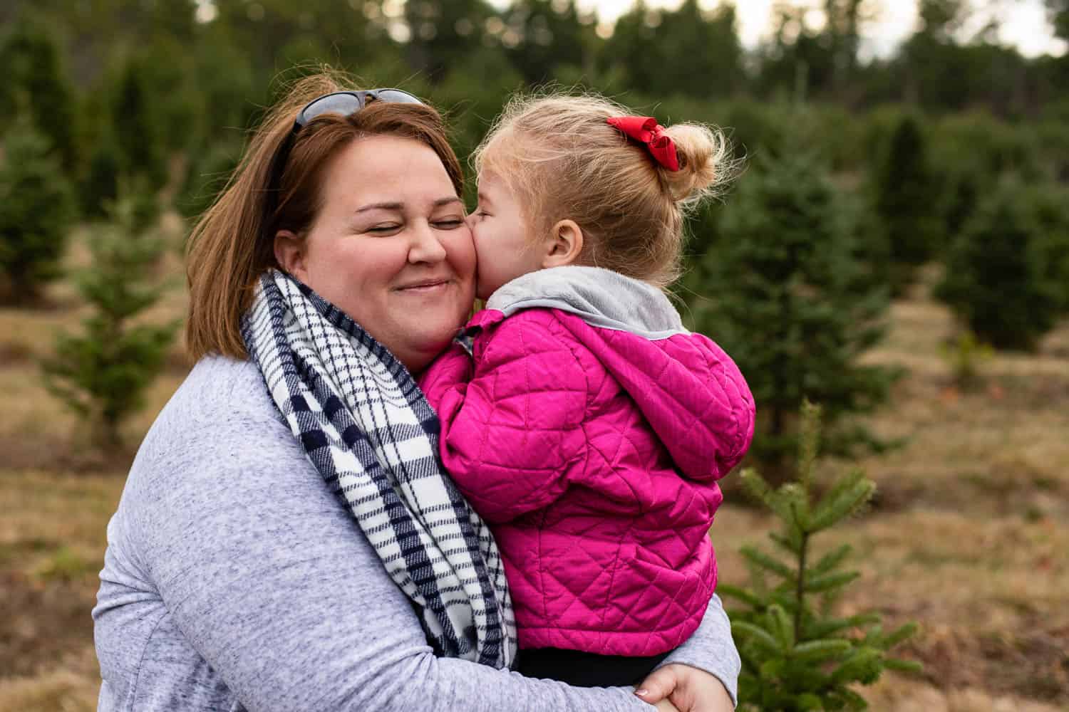 A young girl in a pink jacket kisses a smiling woman on the cheek while they stand outdoors among evergreen trees in Maine.