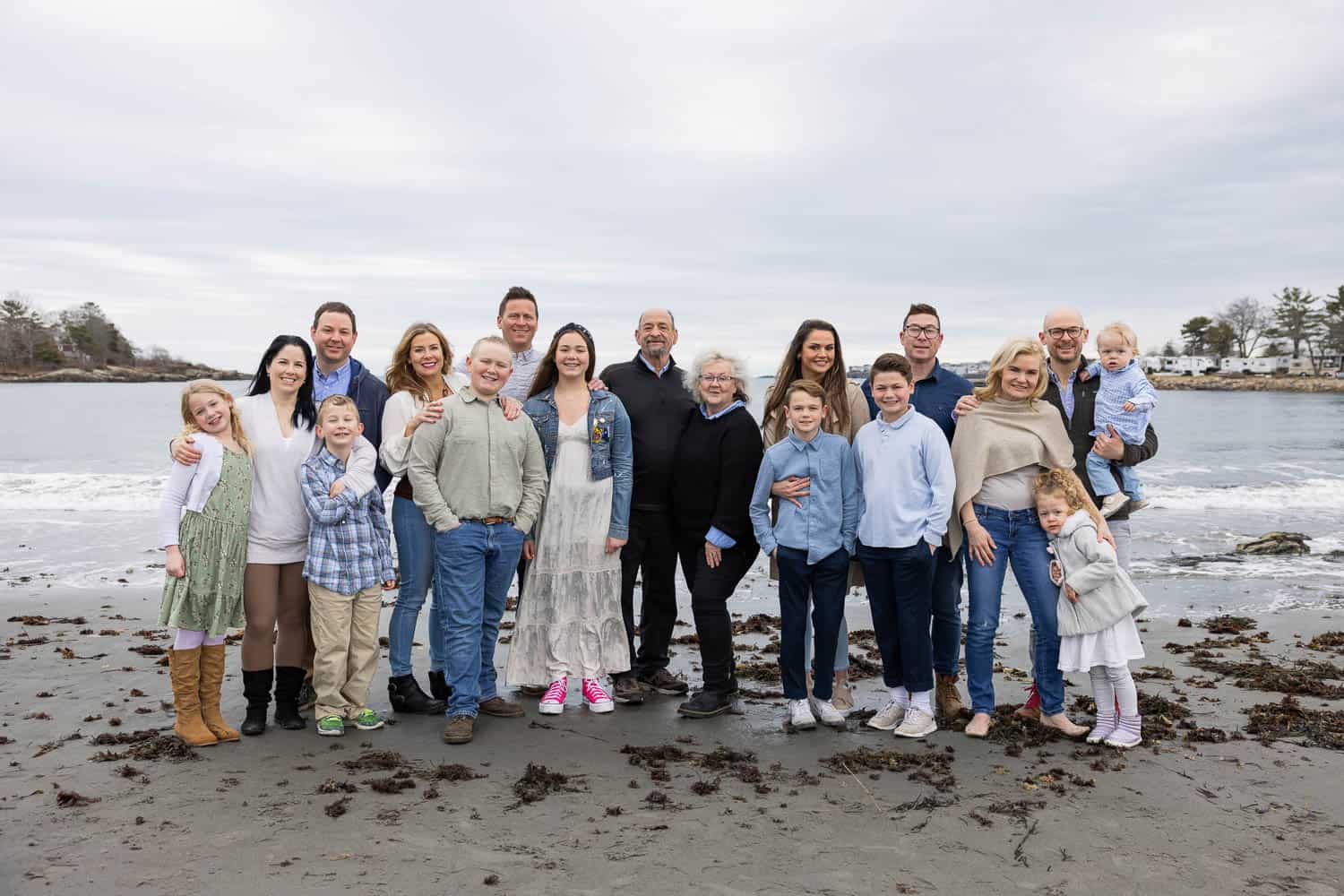 A large family group of adults and children stands together on a beach with the ocean and cloudy sky in the background.