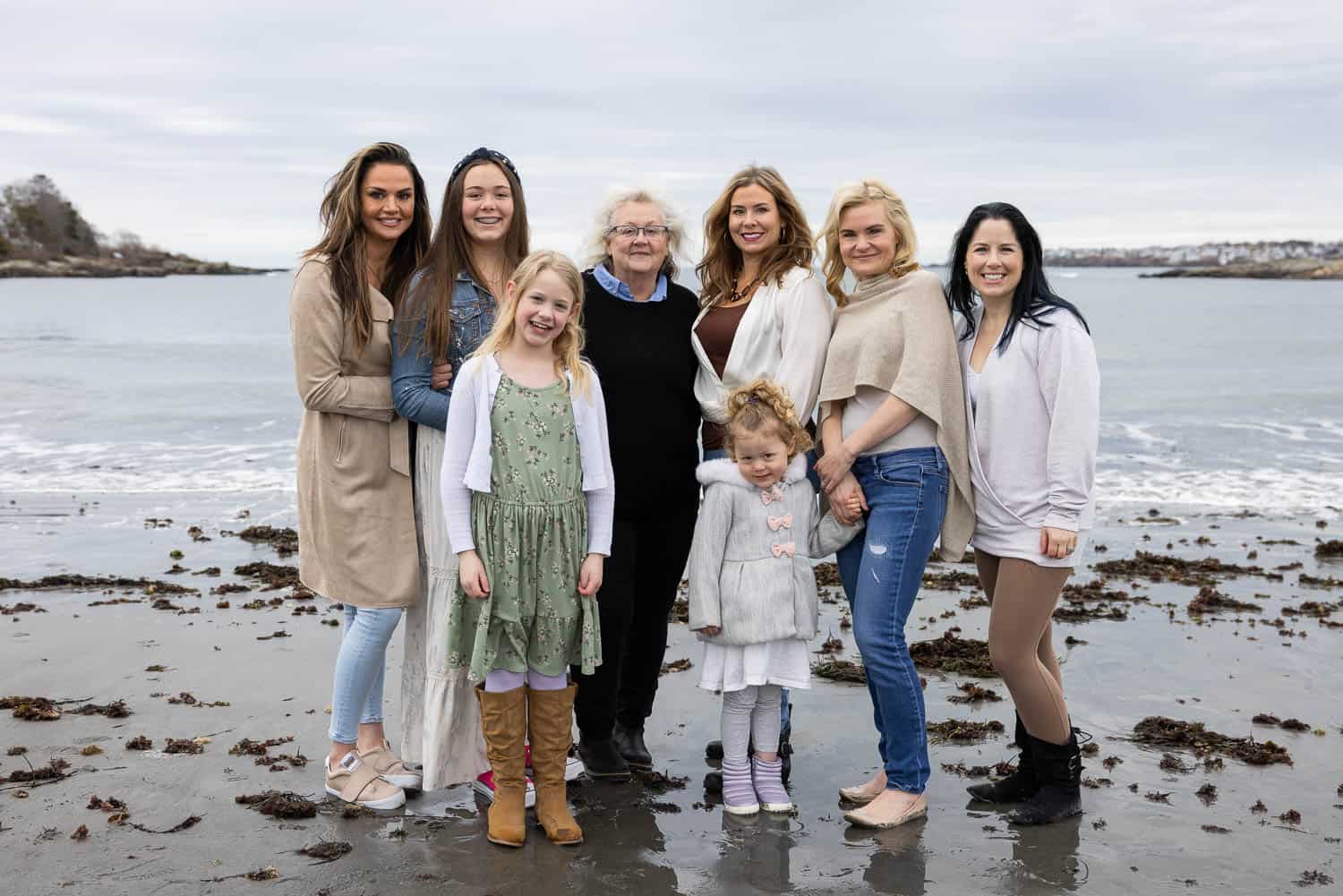 Six women and two young girls stand together on a beach with the ocean and cloudy sky in the background in Maine.
