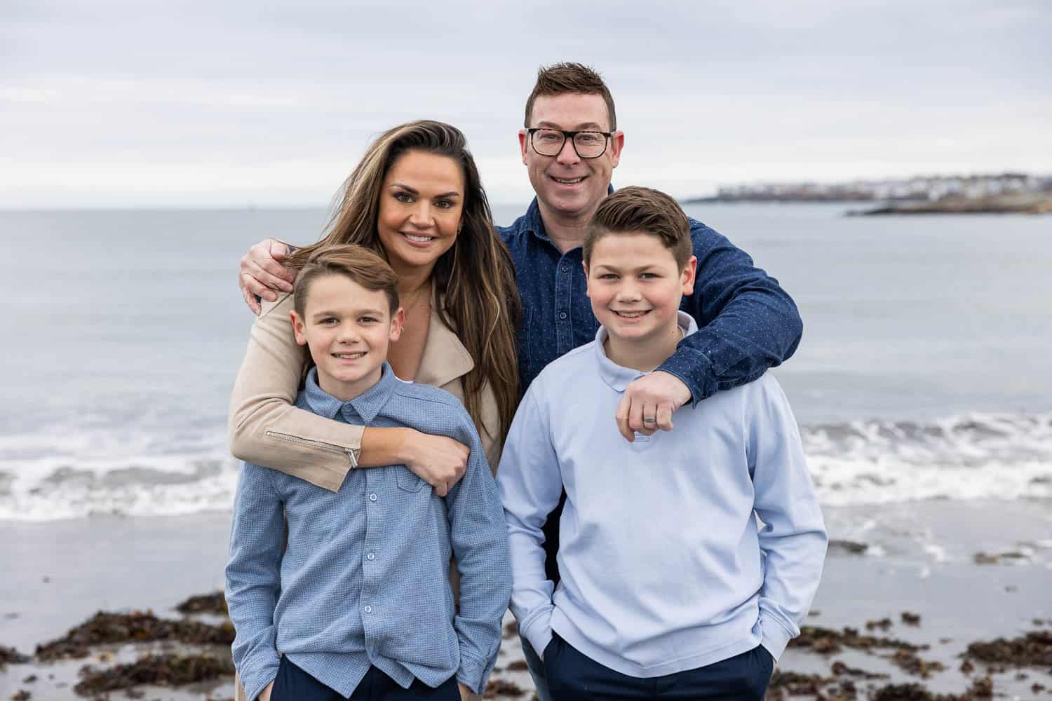 Four people, two adults and two children, stand together on a beach with the ocean and cloudy sky in the background.
