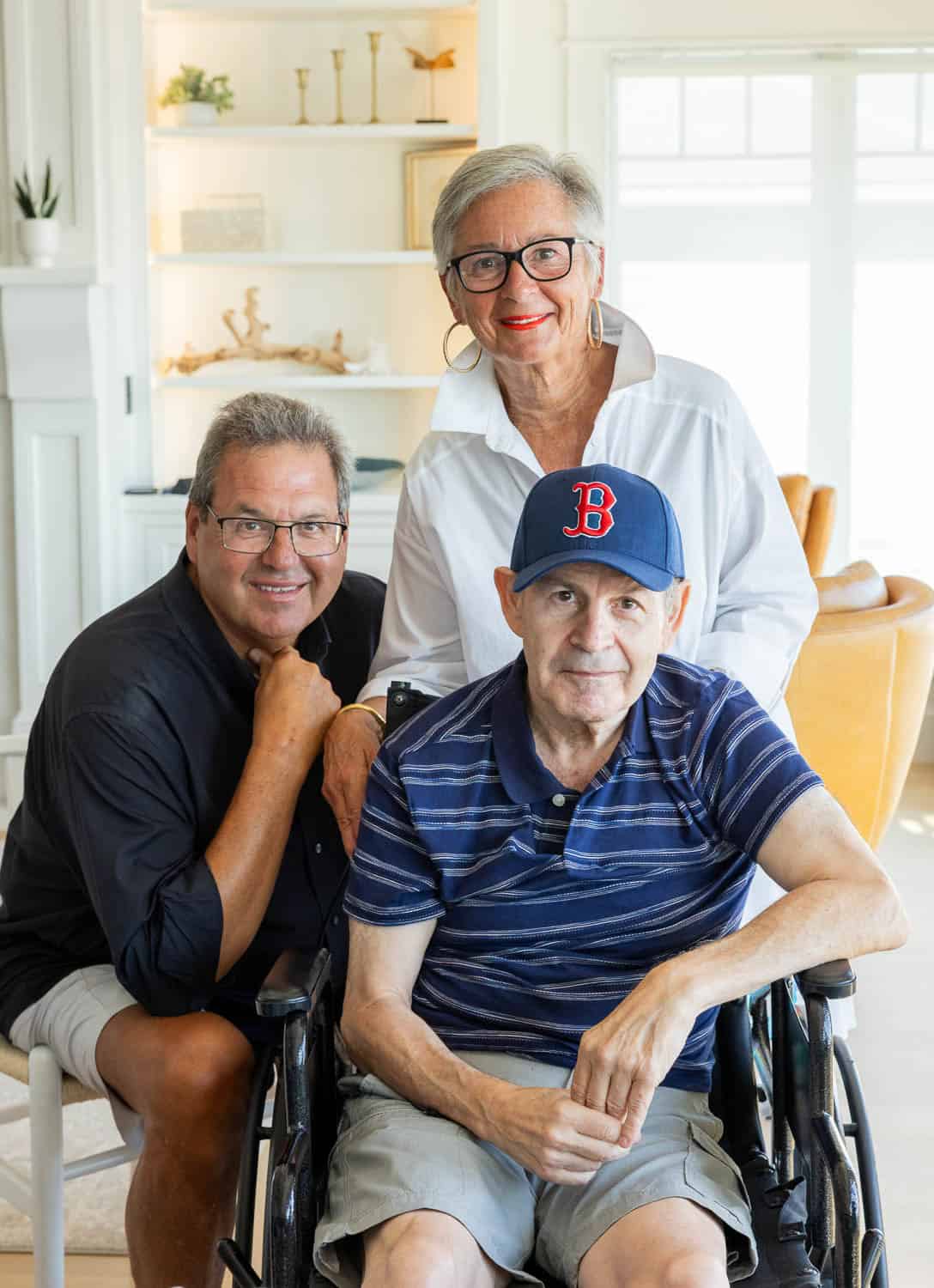 Three adults indoors; one man sits in a wheelchair wearing a Boston Red Sox cap, while another man and a woman stand behind him, all smiling at the camera.