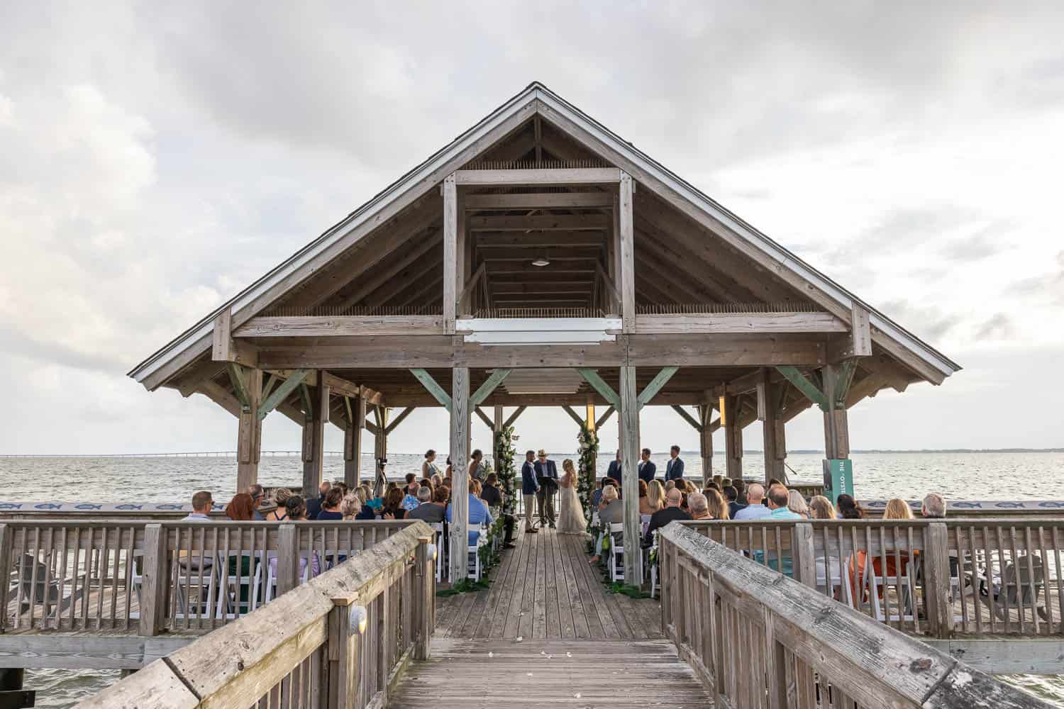 A wedding ceremony takes place under a wooden pavilion on a dock overlooking the water, with guests seated and the sky cloudy above.