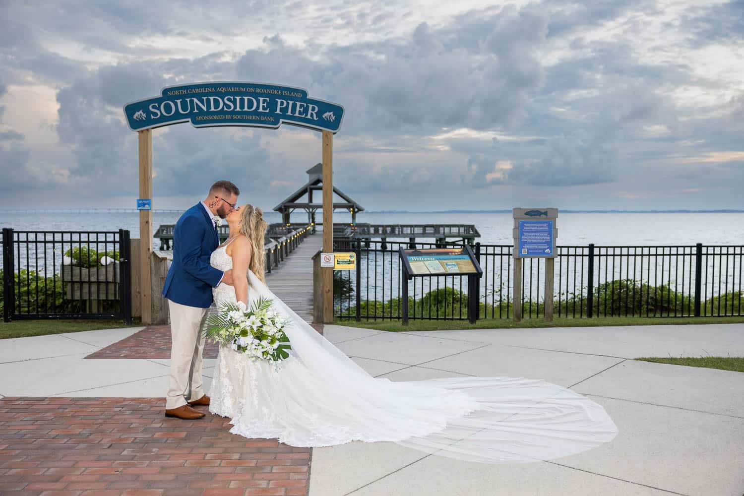 Bride and groom stand at the Soundside Pier entrance with water and a cloudy sky behind them. She holds a bouquet and wears a long white dress.