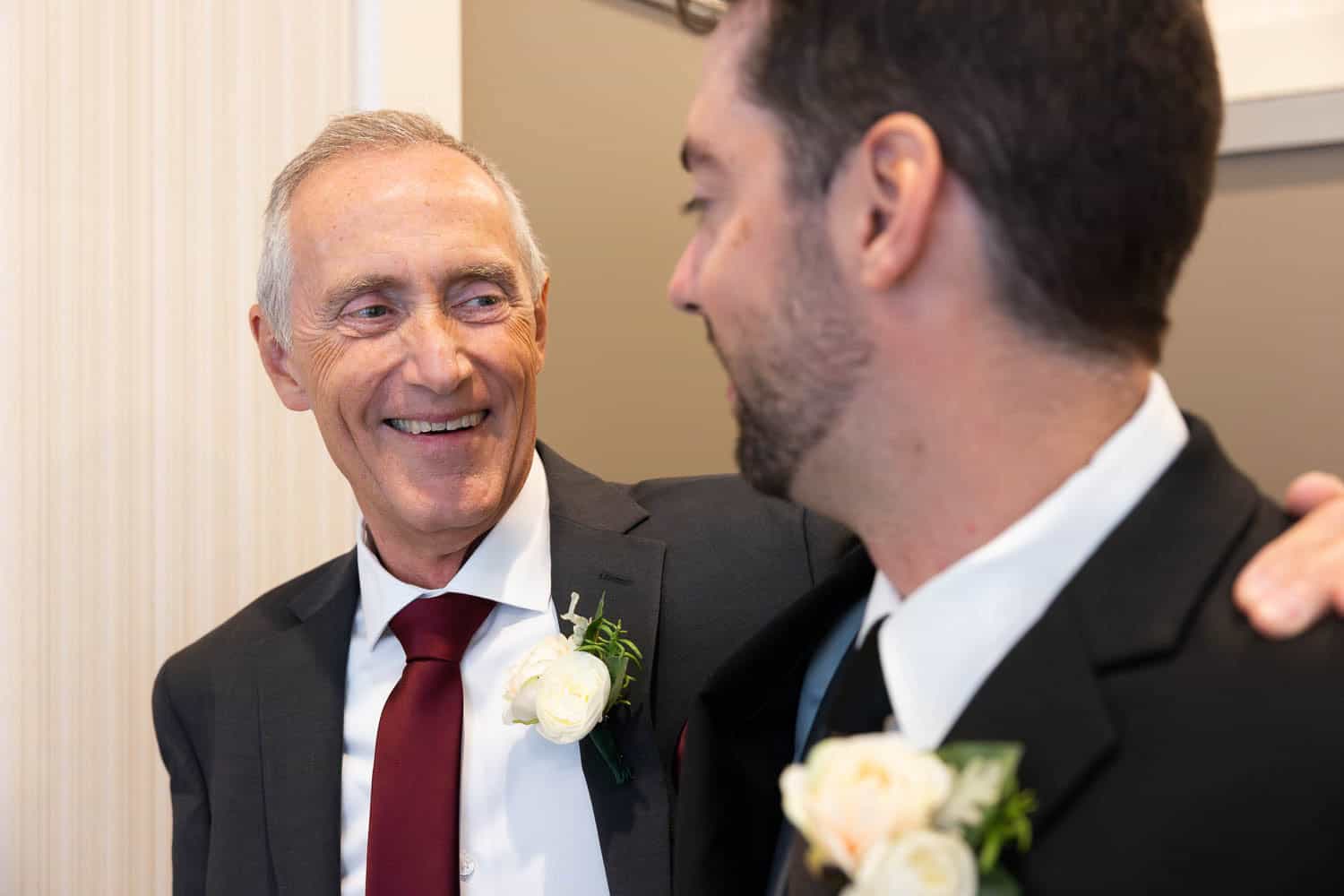 Two men in suits with boutonnieres smile at each other indoors, suggesting a celebratory event such as a wedding.