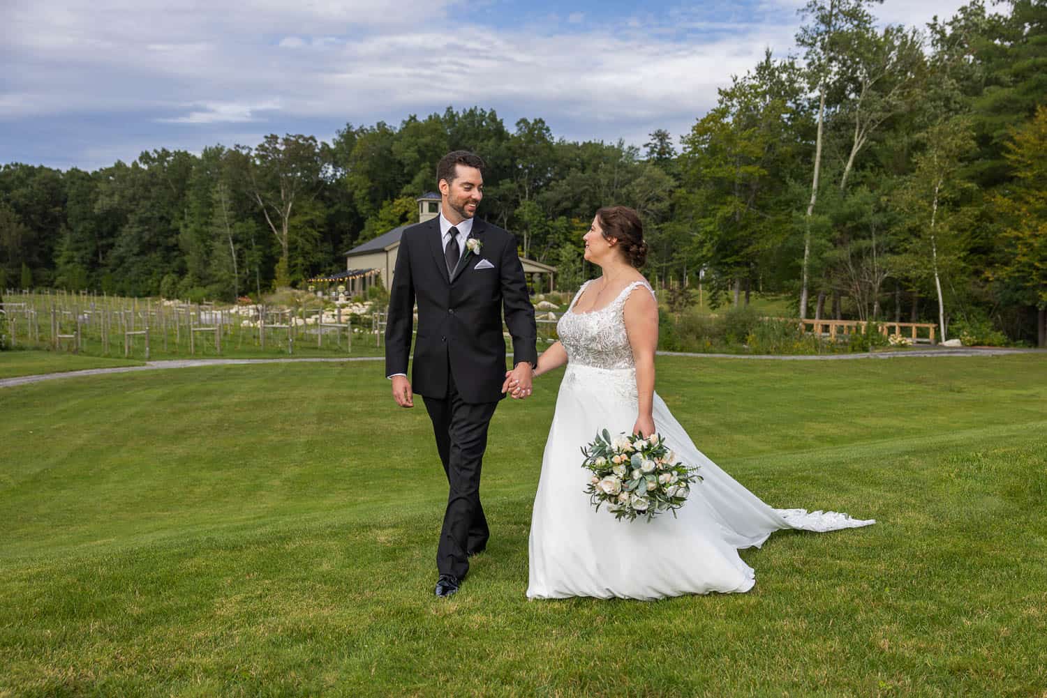 A bride and groom walk hand in hand on a grassy lawn, with trees and a building in the background. The bride holds a bouquet of flowers.