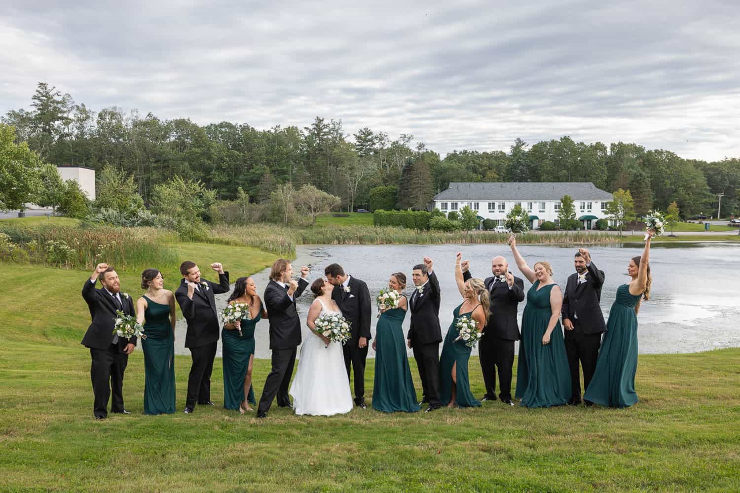 A wedding party outdoors near a pond, with the bride and groom kissing in the center, surrounded by bridesmaids in green dresses and groomsmen in black suits.