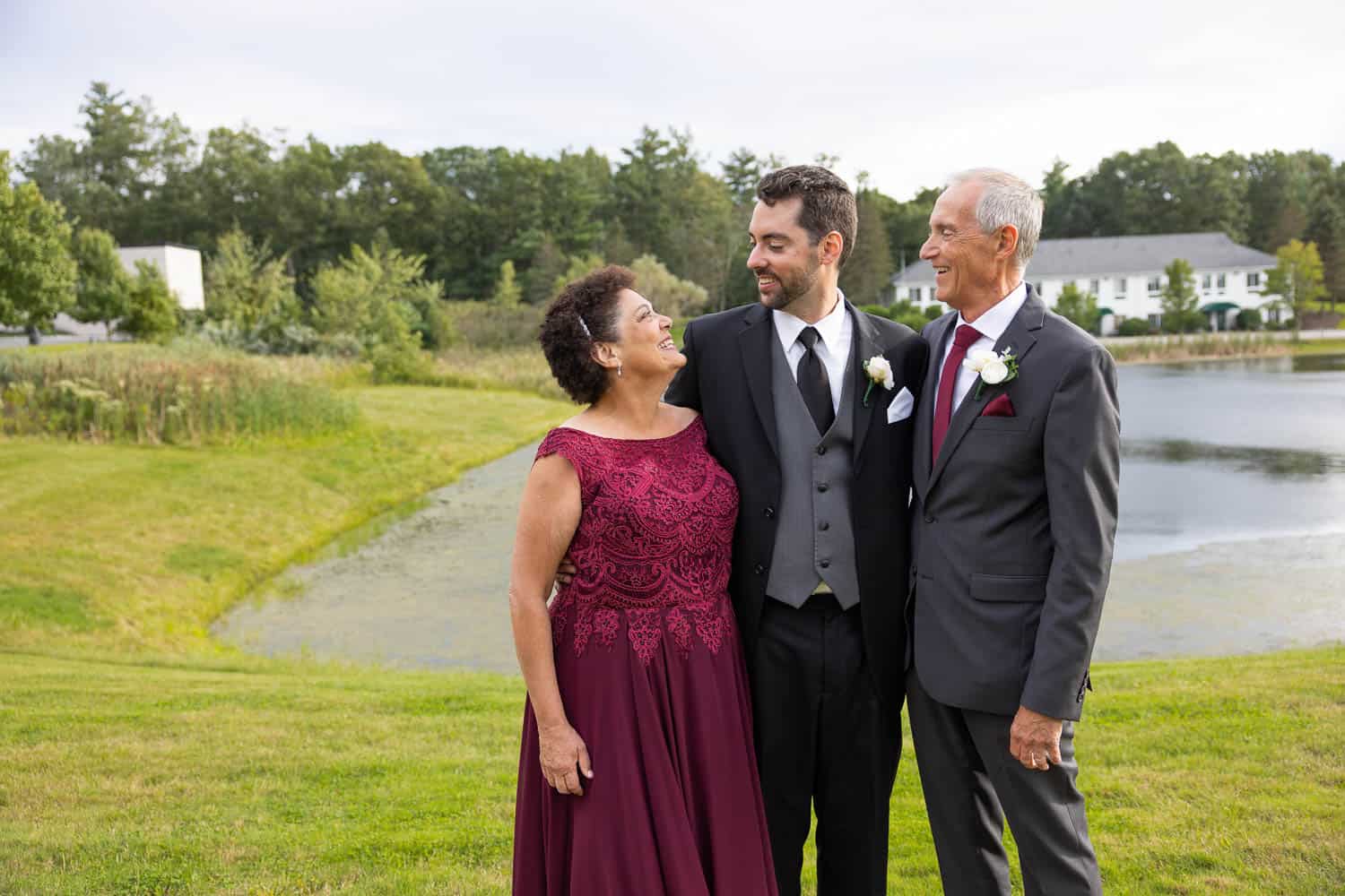 A man in a suit stands between an older woman in a burgundy dress and an older man in a gray suit by a pond, all smiling at each other.