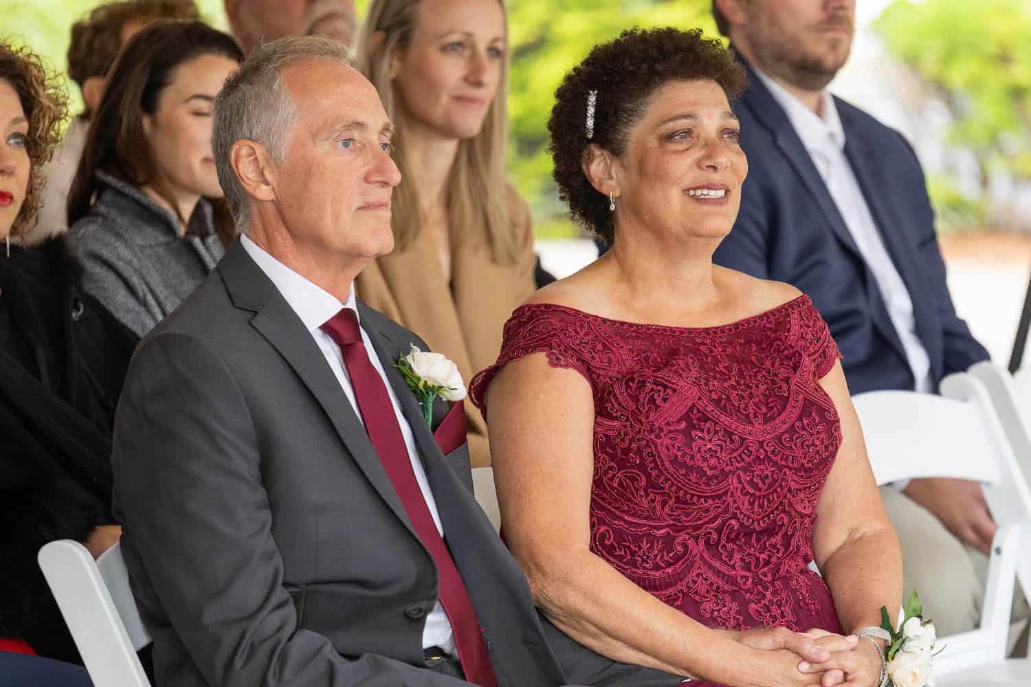 A man in a suit and a woman in a red dress sit side by side on white chairs at an outdoor event, with other attendees seated behind them.