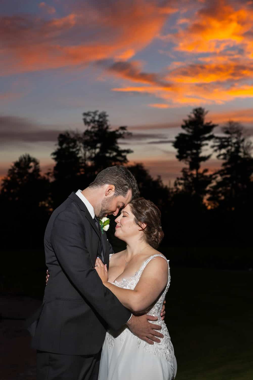 A bride and groom embrace outdoors at sunset, with trees silhouetted against a colorful sky.