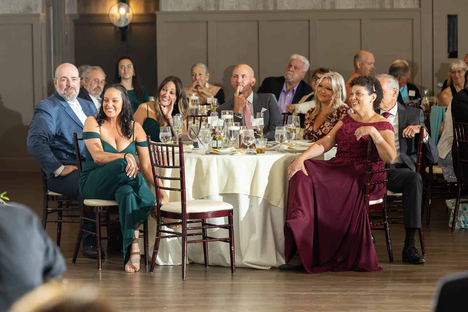 A group of people in formal attire sit around a table at an wedding reception, attentively watching something off-camera.