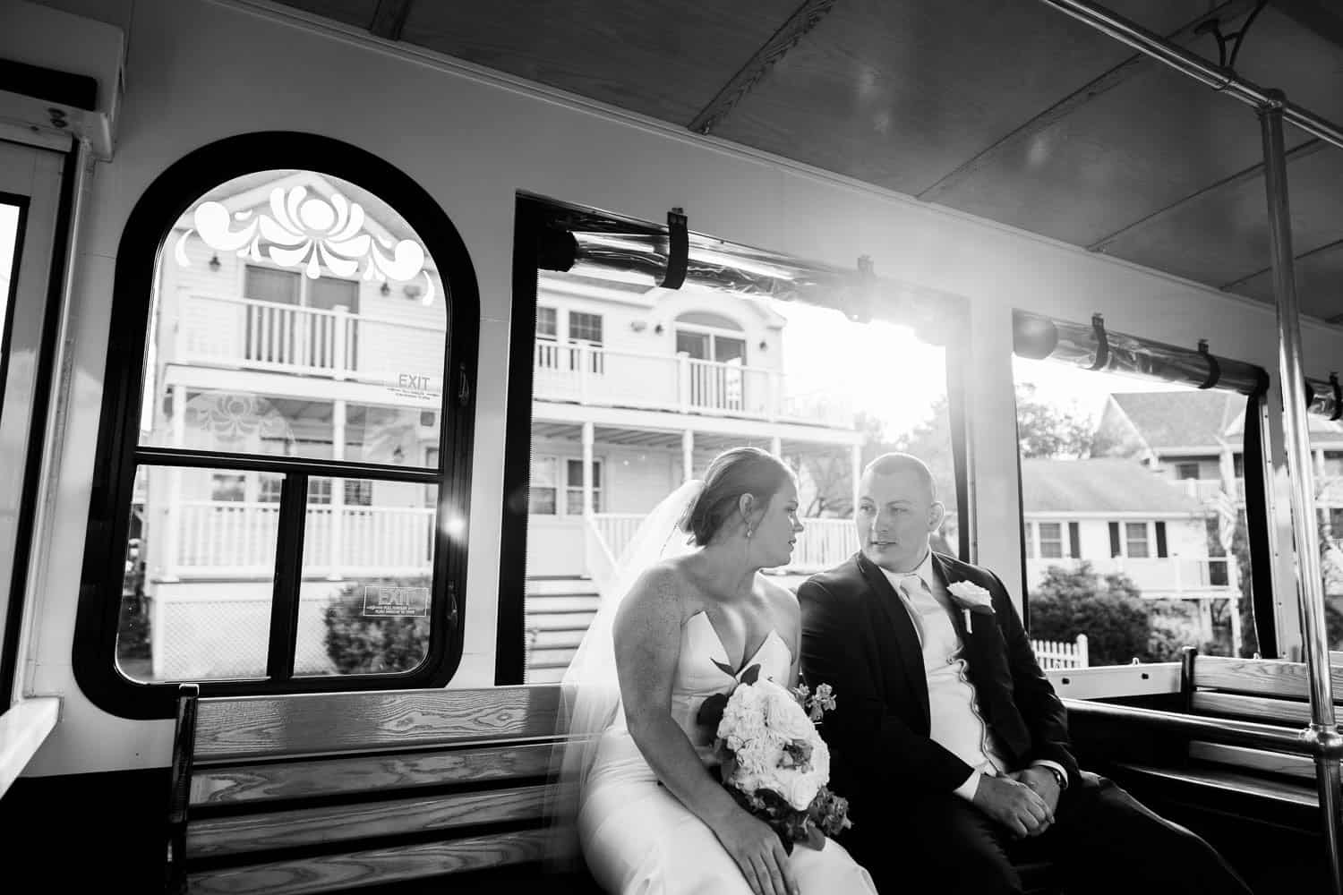 A bride and groom sit together on a trolley, facing each other. The bride holds a bouquet, and sunlight streams through the window behind them.
