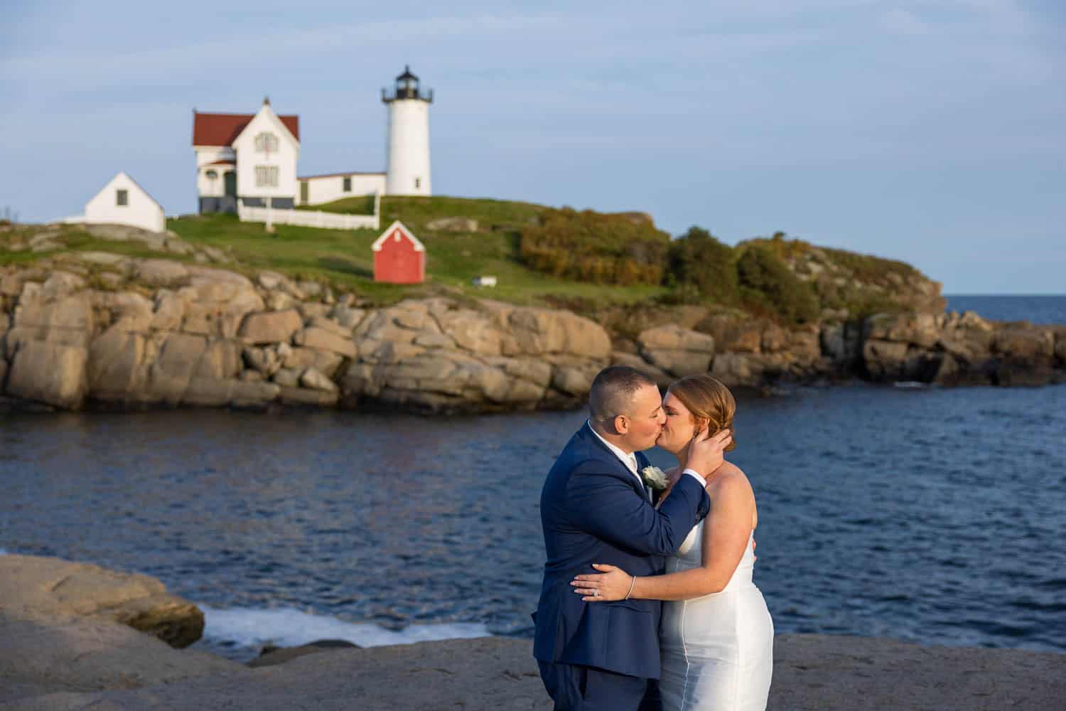 A bride and groom kiss on a rocky Maine shoreline with a lighthouse, white house, and small red building on a grassy hill in the background.