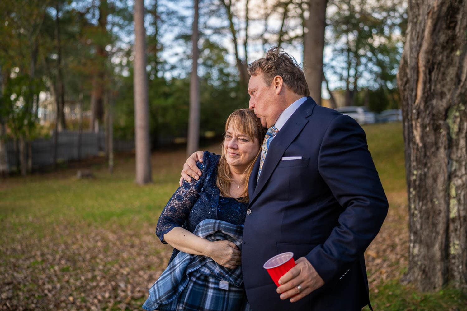 A man in a suit hugs and kisses a woman on the forehead outdoors; she is emotional and holding a plaid blanket. Trees and fallen leaves are in the background.