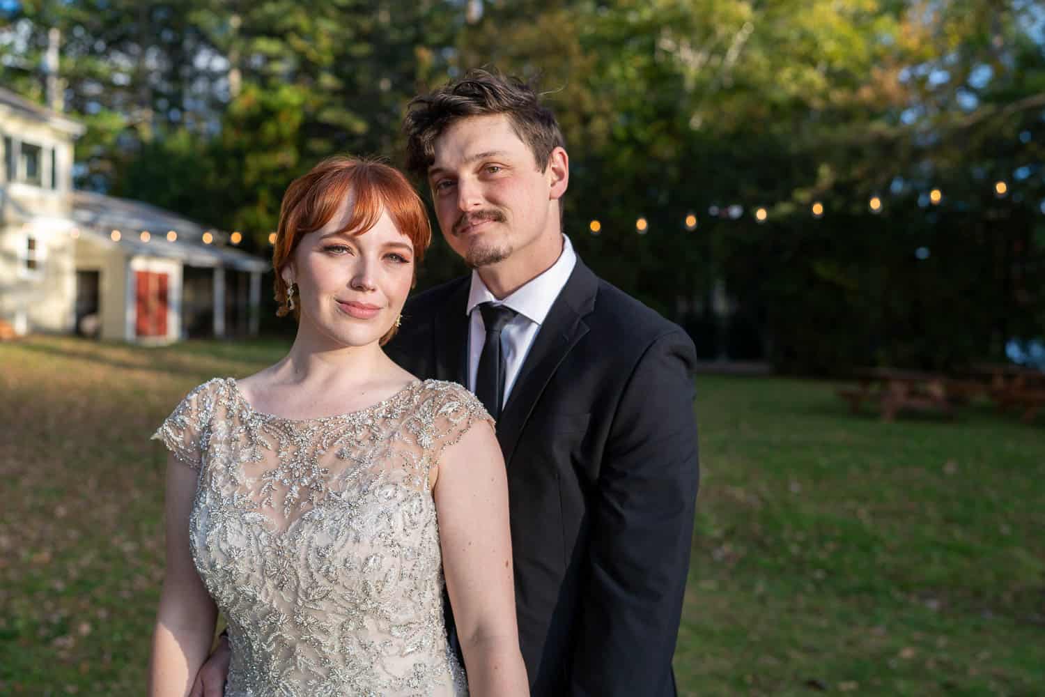 A formally dressed couple stands close outdoors; the woman in a beaded white dress, the man in a black suit and tie. String lights and trees in the background.