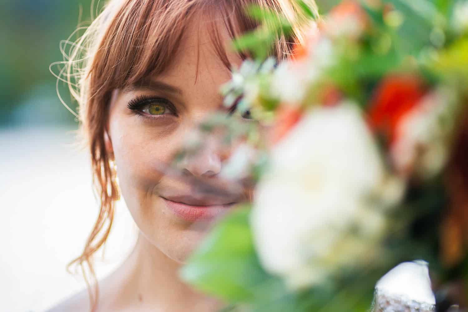A woman with red hair smiles softly, with part of her face partially obscured by a colorful bouquet of flowers in the foreground.