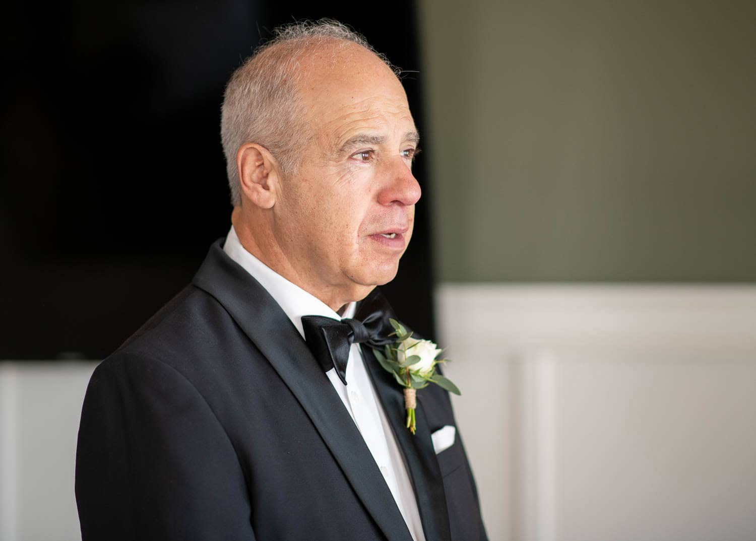 A man in a black tuxedo with a bow tie and boutonnière stands indoors, looking to the side.