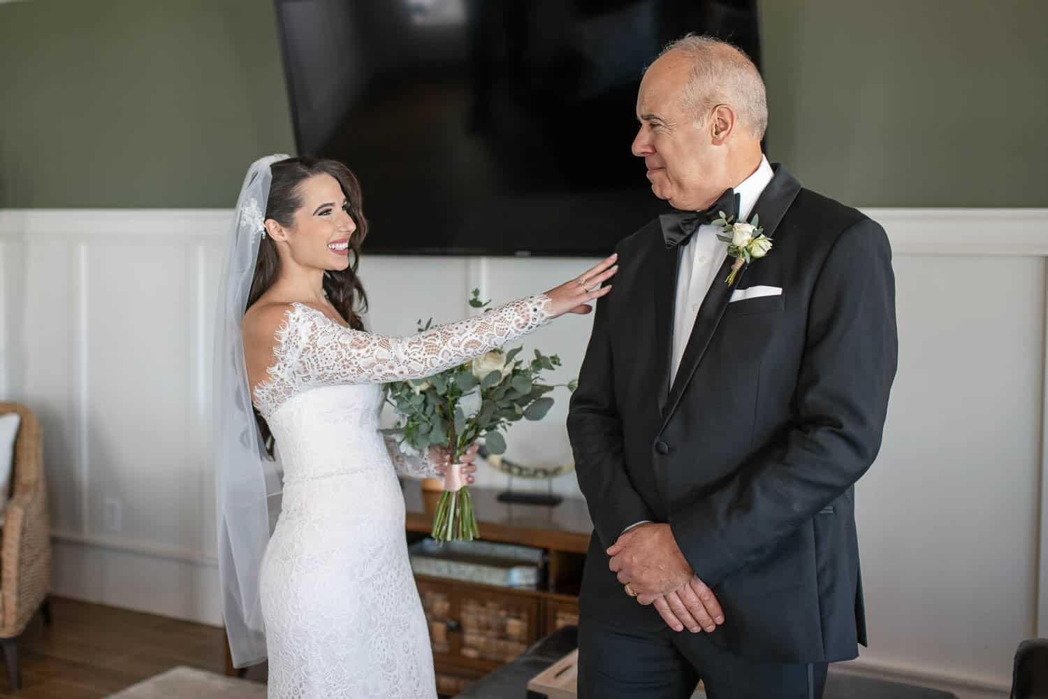 A bride in a white dress, holding a bouquet, smiles and reaches toward an older man in a tuxedo, who looks at her.