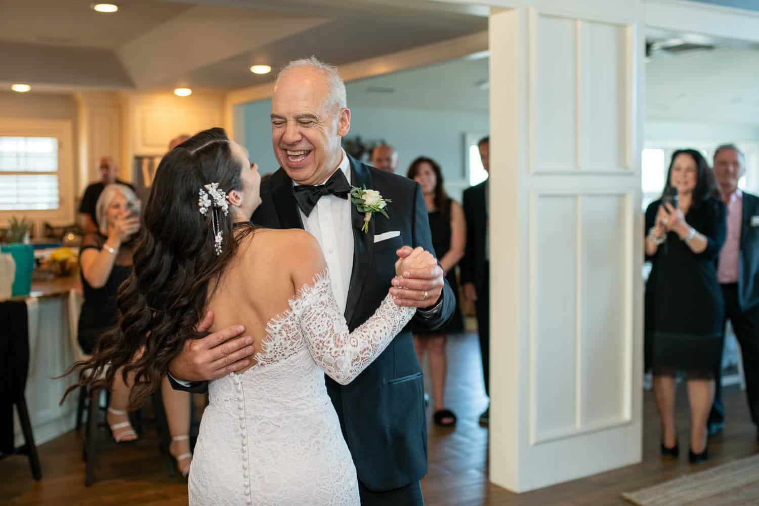 A bride in a white dress dances with an older man in a suit and boutonnière at a wedding reception while guests watch and take photos.