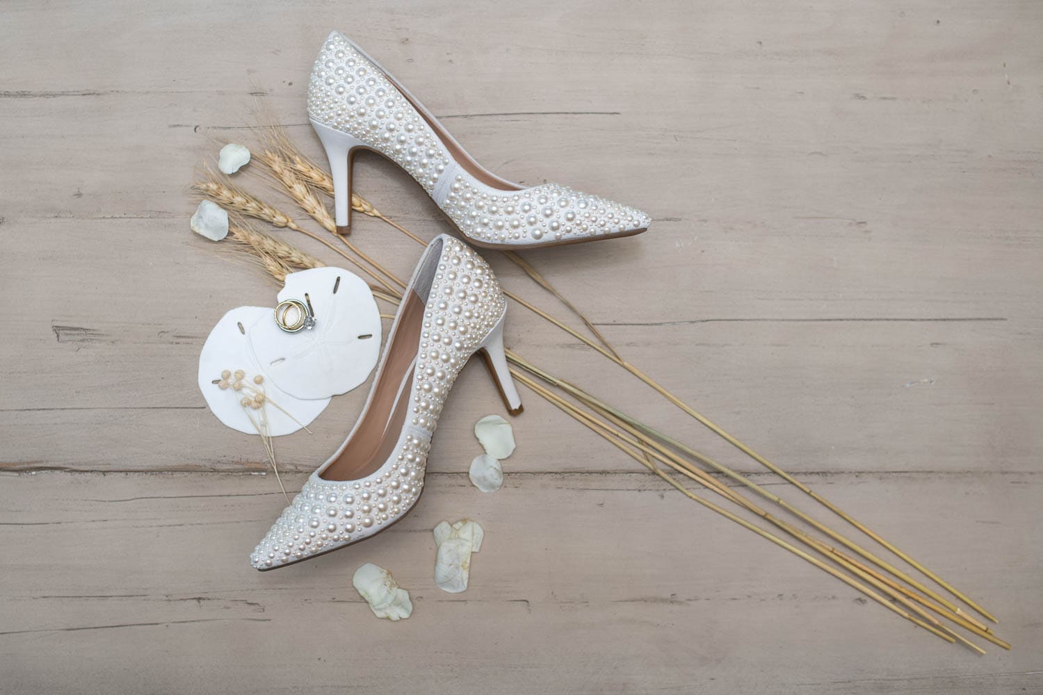 Two white high-heeled shoes with pearl detailing are placed on a wooden surface, alongside wheat stalks, sand dollars, white rose petals, and two gold rings.