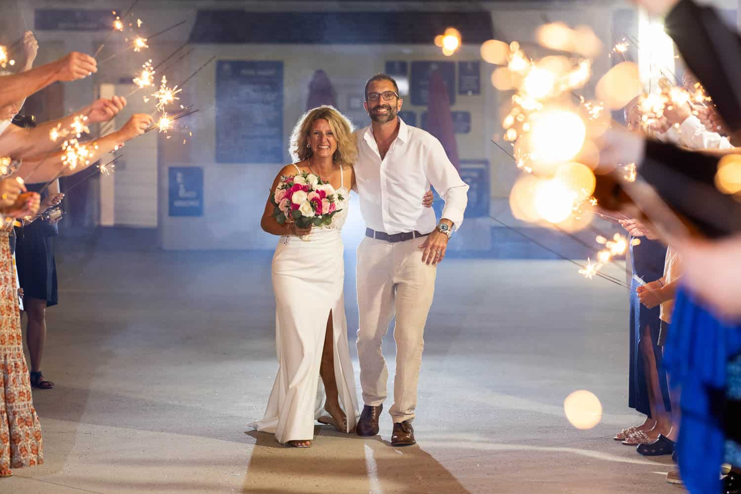 Questions to Ask a Wedding Photographer A couple in white attire walks together while holding flowers, surrounded by people holding sparklers in a festive indoor setting.