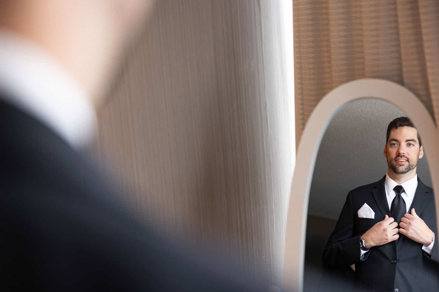 A man in a suit adjusts his jacket while looking at his reflection in a mirror.