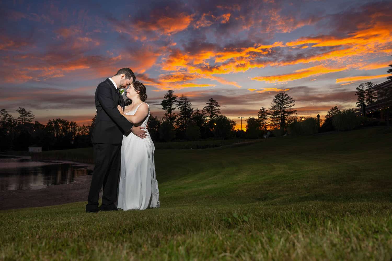 A couple in formal attire embraces on a grassy hill during sunset, with a colorful sky and silhouetted trees in the background.