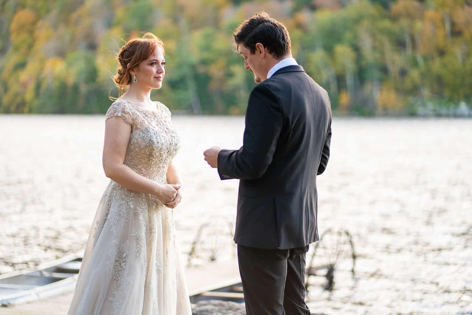 A bridal couple stands near a lake. The woman is in an ornate dress, and the man is in a suit. They are facing each other, trees in the background.