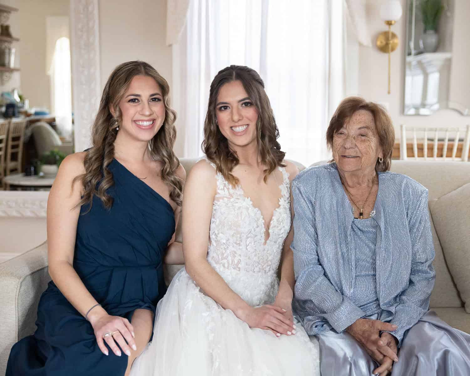 Three women sitting on a couch; the middle one is in a wedding dress, with women in a dark dress on the left and an elderly woman in a light outfit on the right.