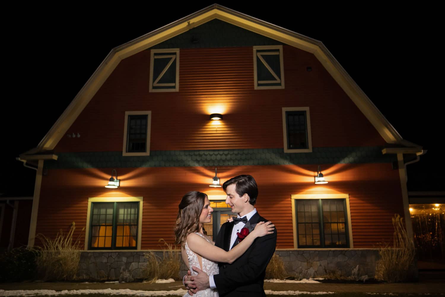 A couple in wedding attire embraces in front of a well-lit, barn-like structure at night.