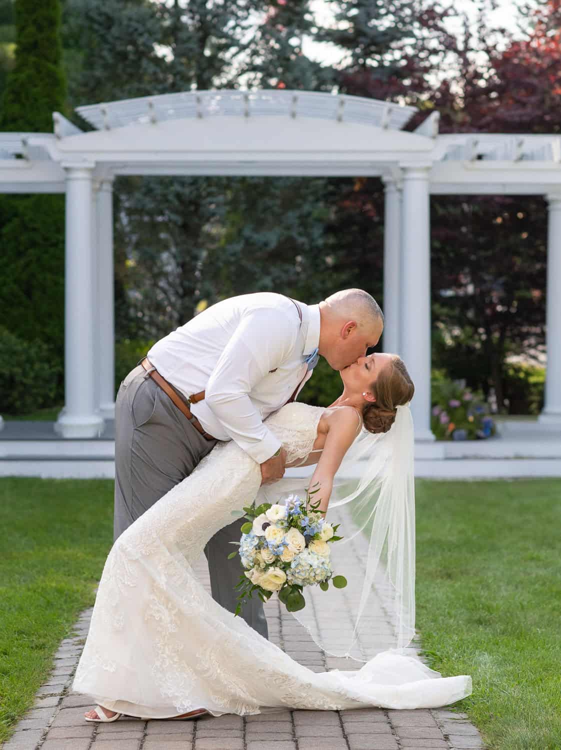 Groom in white shirt dips bride in lace gown and veil for a kiss; she holds a bouquet. Background features a white pergola and greenery.