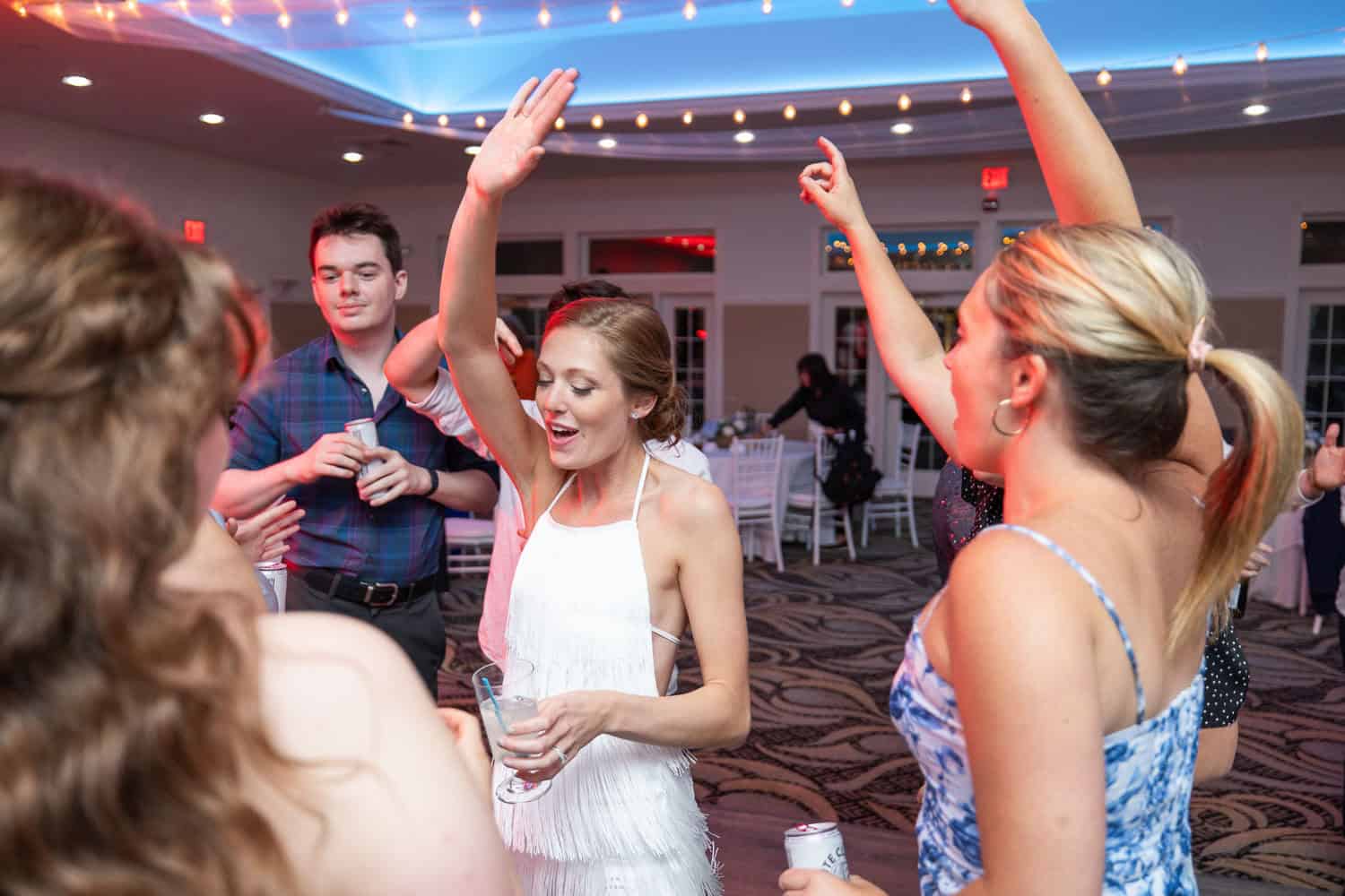 People are dancing and smiling in a brightly lit room. A woman in a white dress holds a drink, raising one hand in the air.