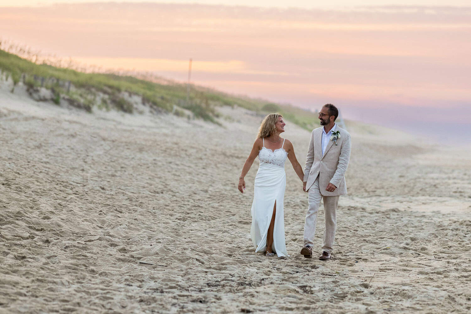 A couple in wedding attire walks hand in hand on a sandy beach at sunset, with grassy dunes in the background.