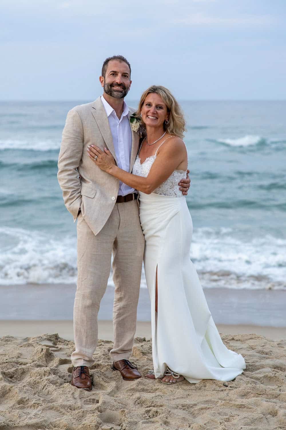 A couple, dressed in wedding attire, stands smiling on a sandy beach with waves in the background.