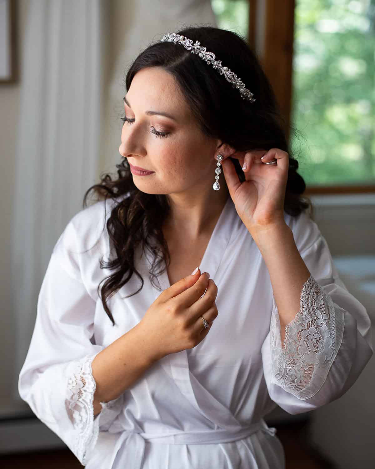 A woman in a white robe adjusts her earring, wearing a jeweled headband. She stands near a window with blurred greenery in the background.