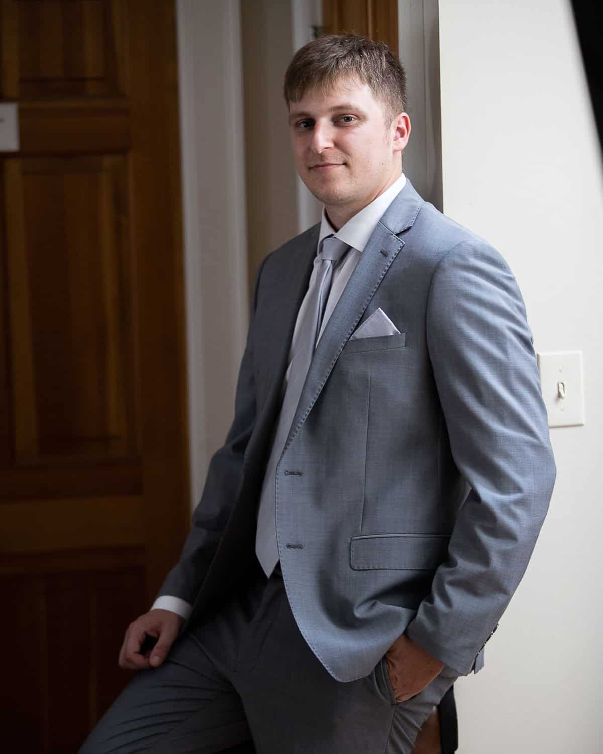Man in a light gray suit leans against a wall, with hands in pockets, near a wooden door.