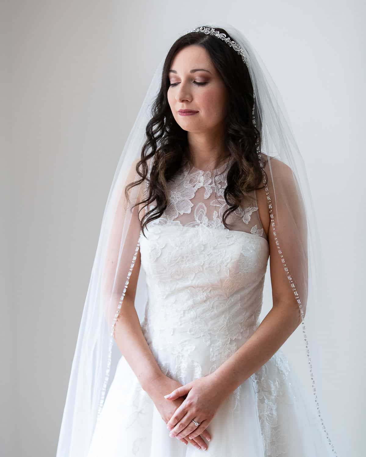 Bride in a white lace wedding dress and veil stands with eyes closed, hands clasped, in front of a plain background.