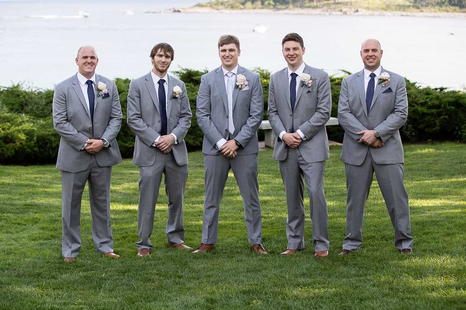 Five men in grey suits stand together on grass, with a body of water and greenery in the background.