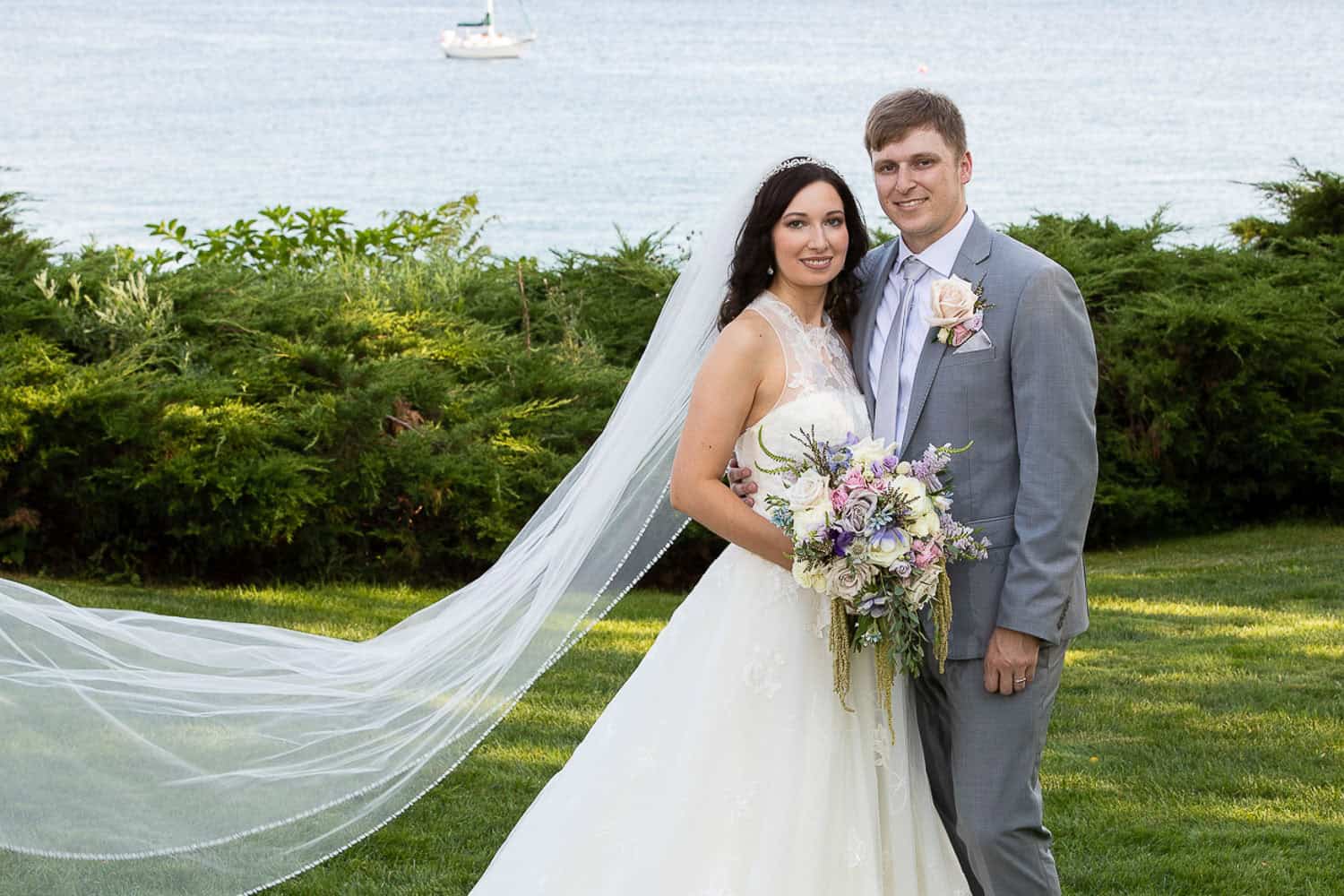 A bride and groom stand together outdoors, with the bride holding a bouquet. The ocean and a sailboat are visible in the background.