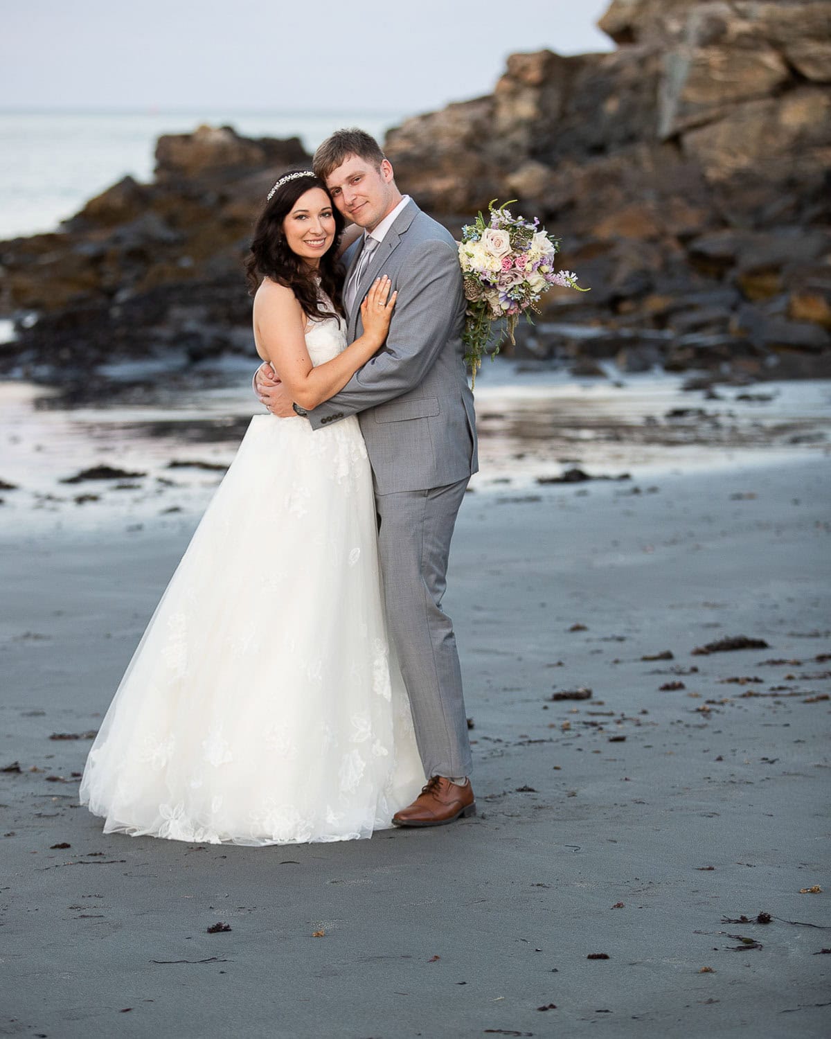 A couple in wedding attire embraces on a sandy beach with rocky background. The bride holds a bouquet and wears a white gown; the groom wears a gray suit.