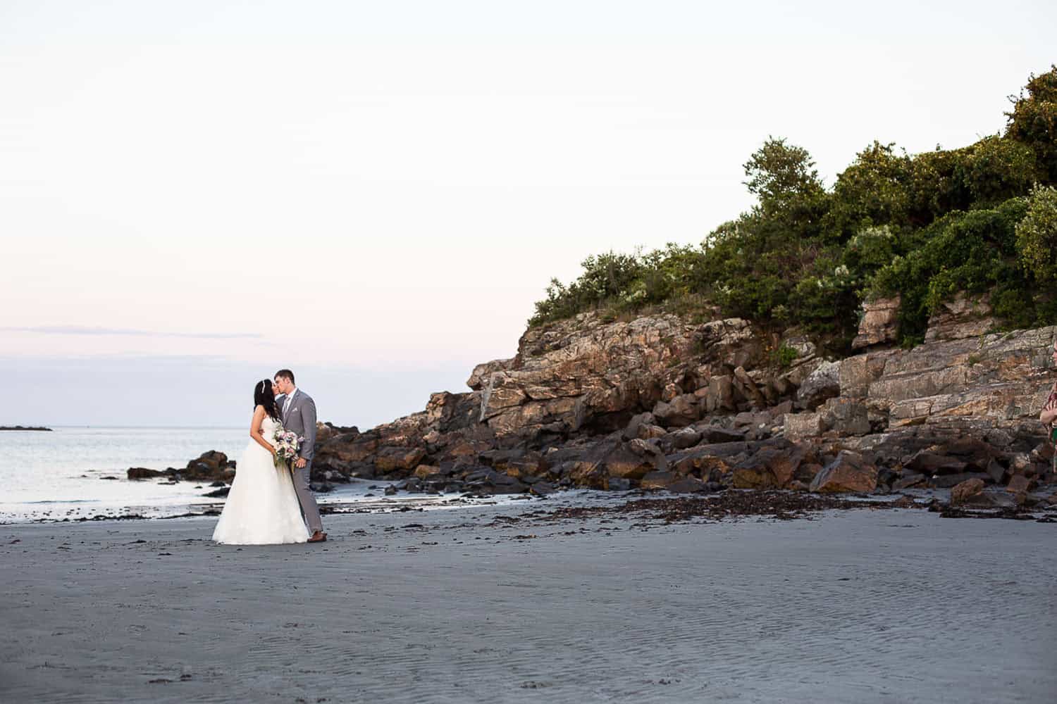 A bride and groom stand on a beach near rocky cliffs, embracing and holding a bouquet, with a calm sea and clear sky in the background.