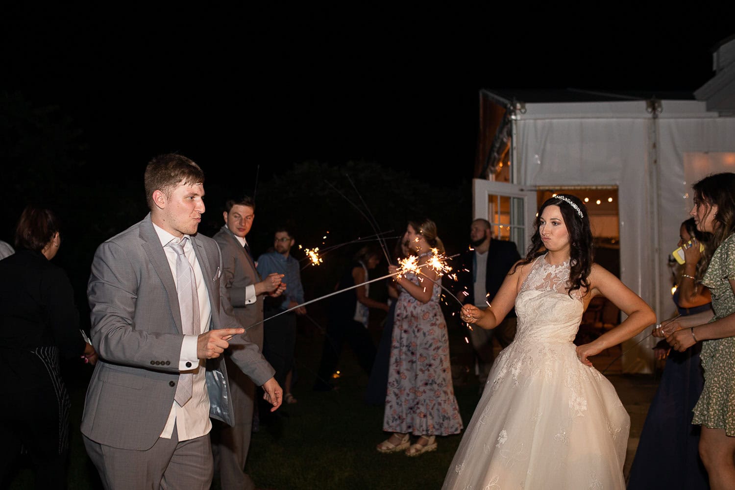 Bride and groom hold sparklers at night surrounded by guests, some with sparklers. A white tent is lit in the background.